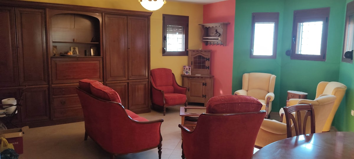 A comfortable living area features a variety of seating options, including two red armchairs and a cream-colored armchair. A wooden cabinet stands against a yellow wall, complemented by green wall accents. Light fills the space from several windows, enhancing the cozy atmosphere.