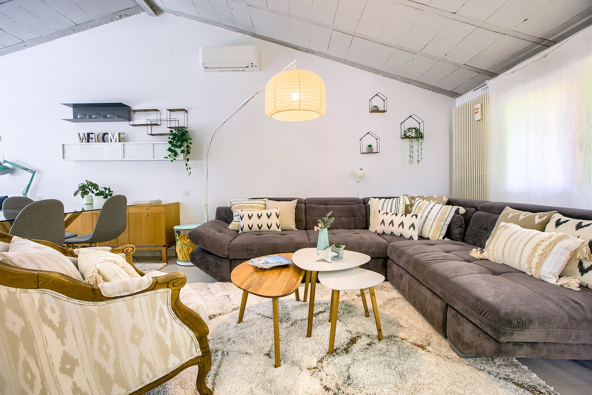 A spacious living area features a comfortable sectional sofa adorned with patterned cushions. Several small tables are arranged on a soft rug, and a floor lamp provides ambient lighting. Decorative wall shelves add character to the bright, inviting space.