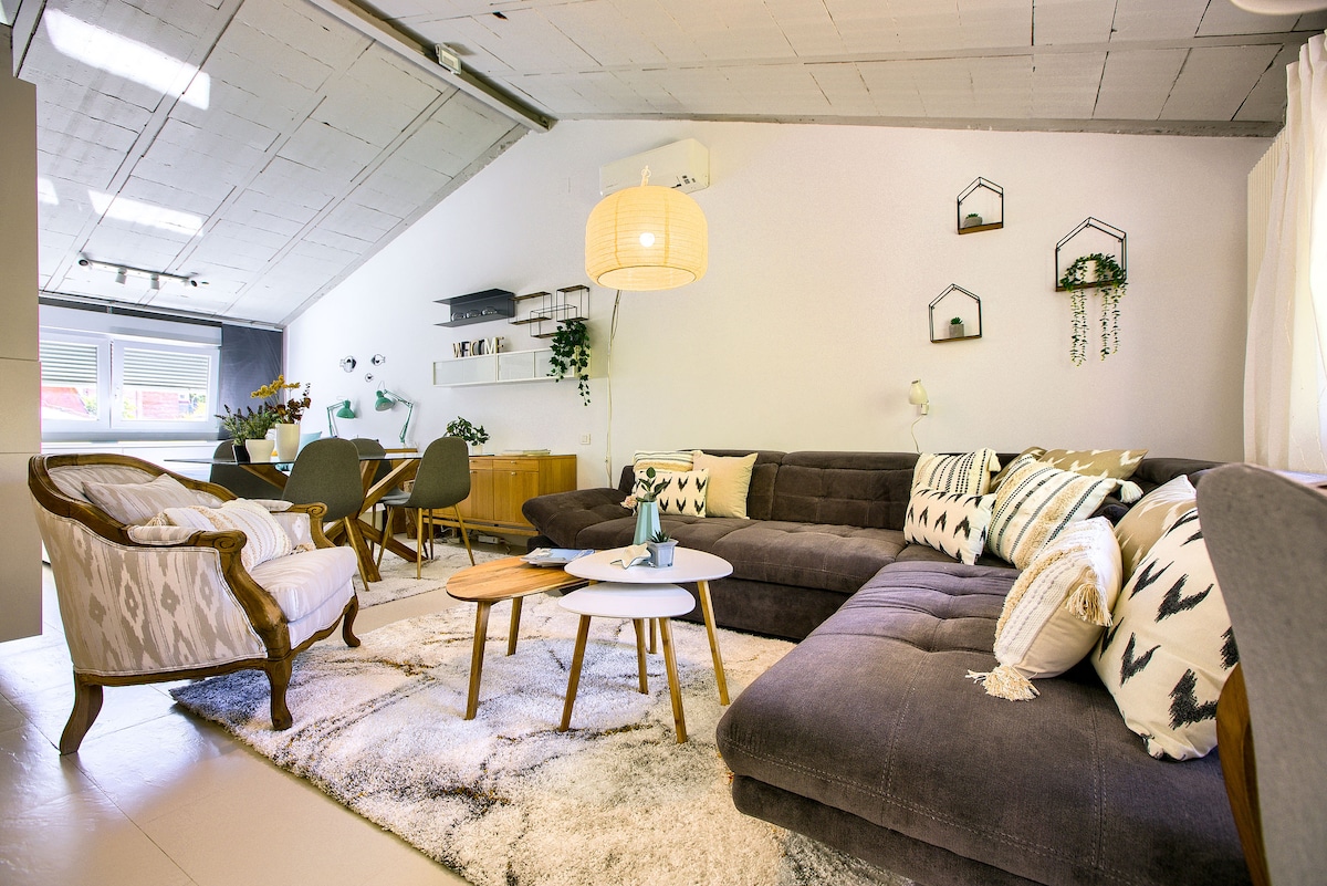 A spacious living area features a dark sectional sofa adorned with patterned cushions and a circular table in the foreground. A cozy armchair and dining table with chairs are visible in the background, complemented by decorative wall shelves and plants.