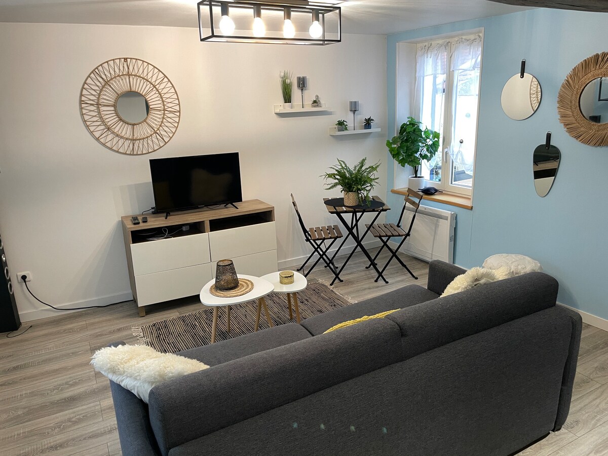 A living area featuring a gray sofa with decorative throws, paired with a small coffee table. A television sits on a console underneath a decorative mirror. Green plants are visible near a window, and a bistro-style table with two chairs enhances the inviting space.