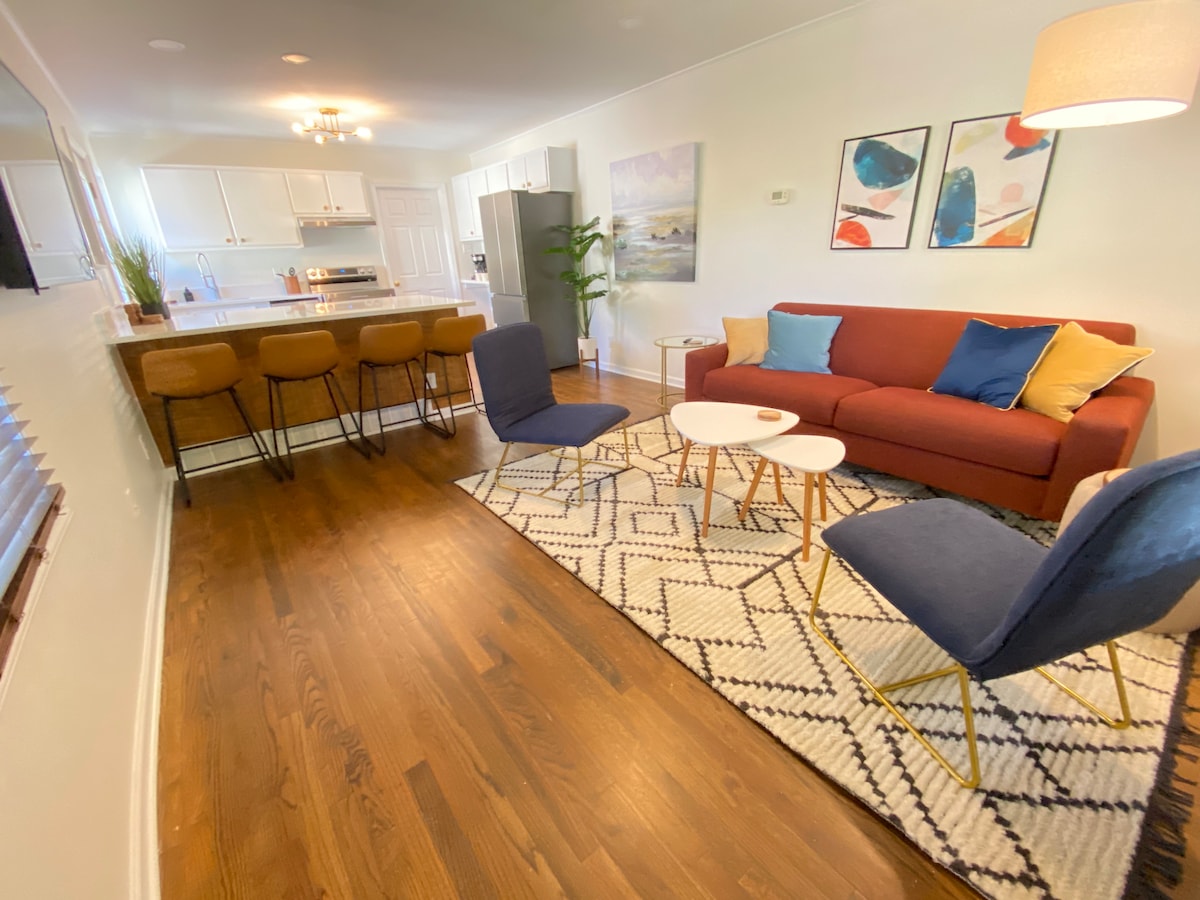 A welcoming living area features a red sofa complemented by colorful throw pillows. Two blue chairs are positioned near a circular coffee table. The space is illuminated by soft ceiling lights, with a modern rug anchoring the wooden floor. A kitchen peninsula with barstools is visible in the background.
