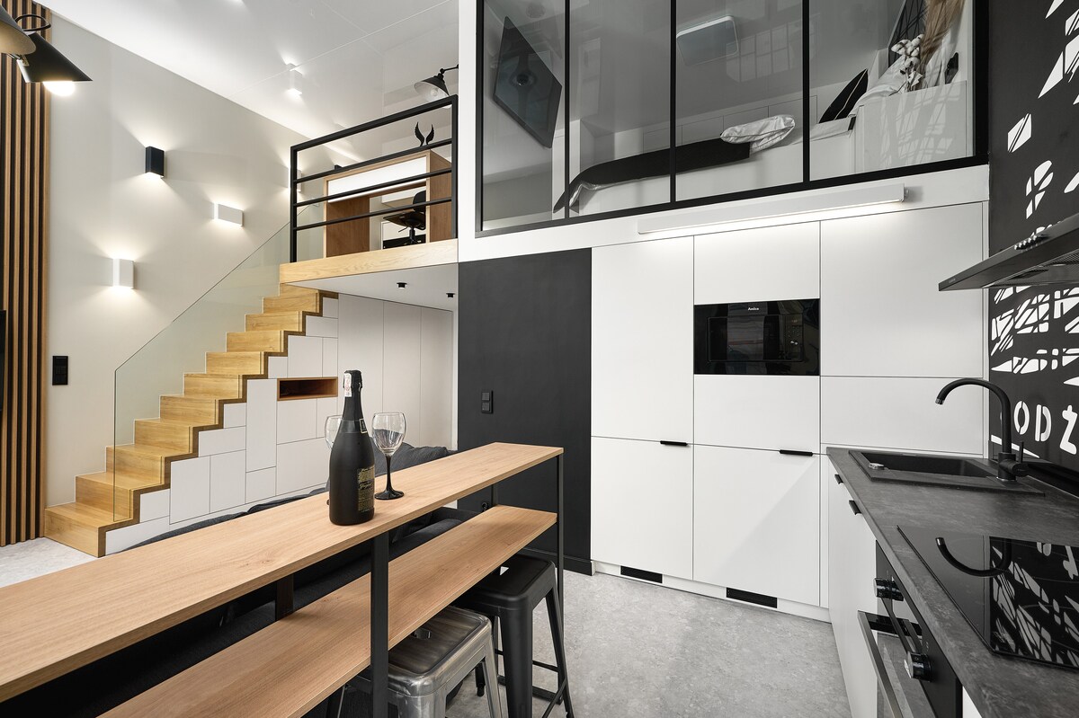 A modern open-concept living space features sleek cabinetry and a dining table with high stools. A stylish staircase leads to a loft area, while light reflects off the minimalist furnishings. The kitchen includes contemporary appliances against a contrasted black wall.