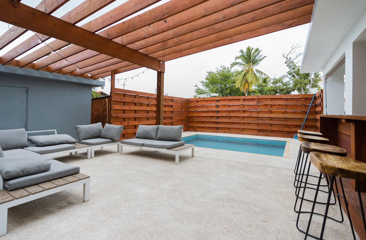 An outdoor area is presented with a small pool surrounded by a wooden deck and a privacy fence. Several lounge chairs with soft cushions are set up for relaxation, while a long bar with high stools offers seating for casual gatherings. Green plants are visible in the background.