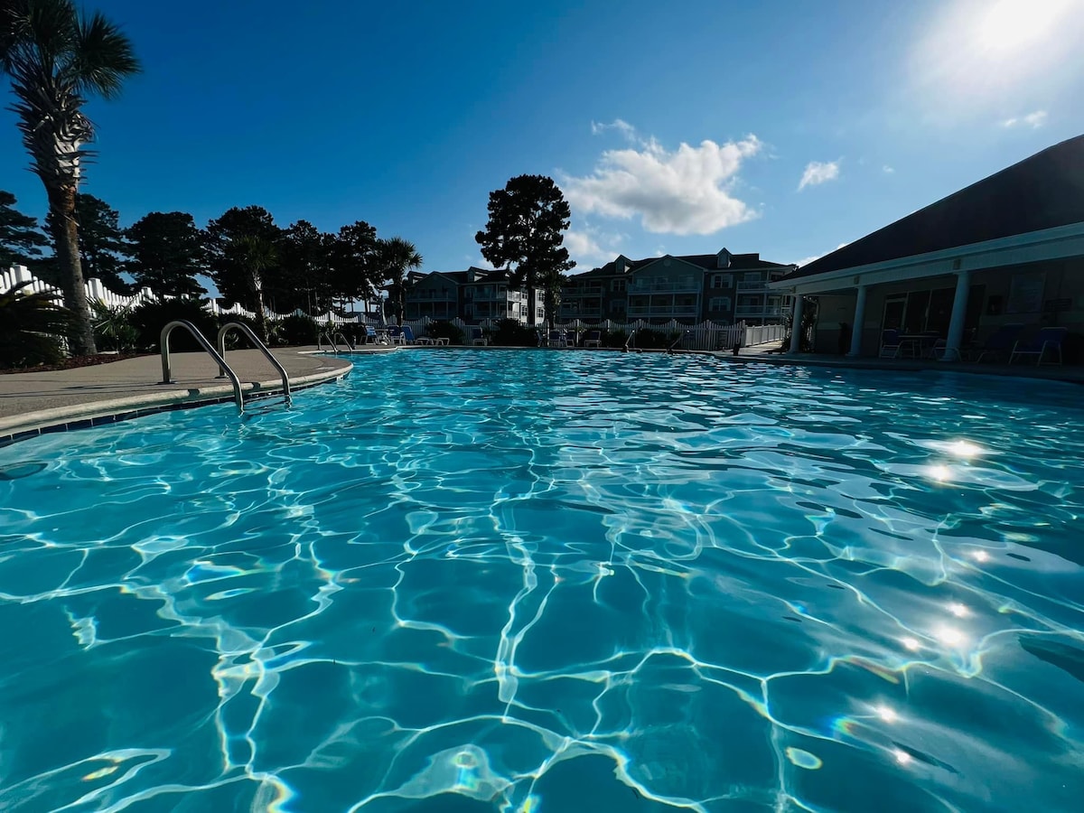 The inground pool is presented with clear blue water, reflecting light in a rippling pattern. Surrounding palm trees and buildings are visible in the background, enhancing the inviting atmosphere of the pool area.