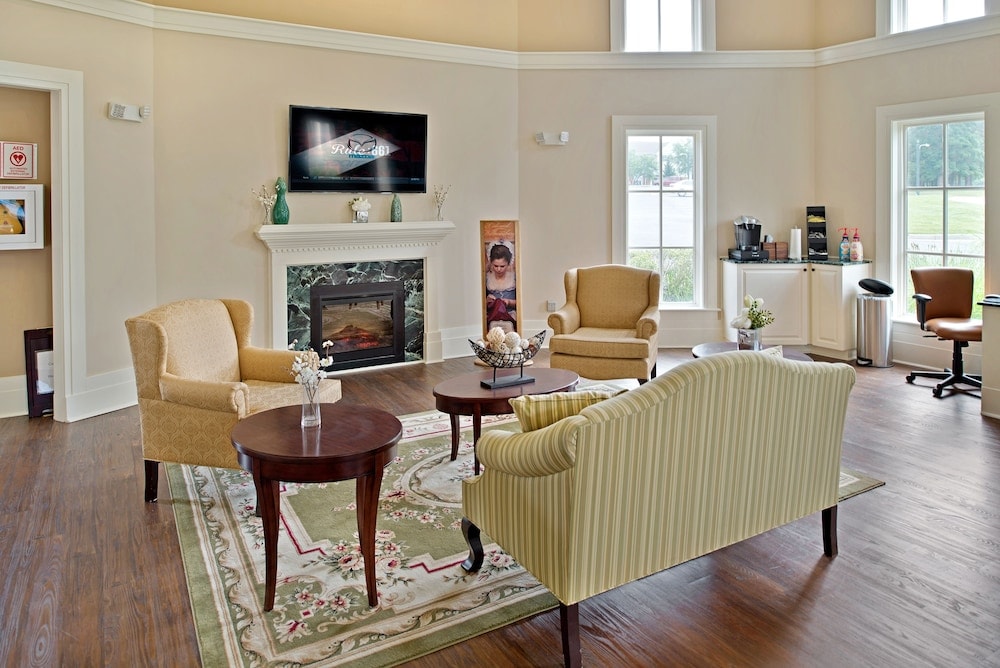 A welcoming common area features a fireplace framed by elegant decoration and a flat-screen TV mounted above. Two plush armchairs and a striped sofa surround a wooden coffee table on a decorative rug, while natural light fills the space through tall windows.