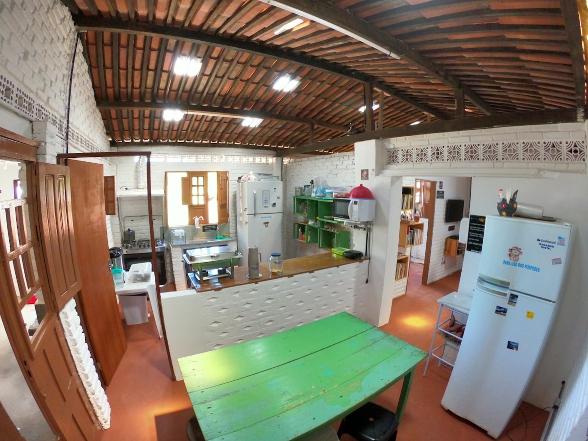 A spacious kitchen features a wooden ceiling with exposed beams, complemented by terracotta tile flooring. A large green table occupies the center, surrounded by appliances and shelves. Natural light filters through the windows, illuminating various cooking areas and a white refrigerator.