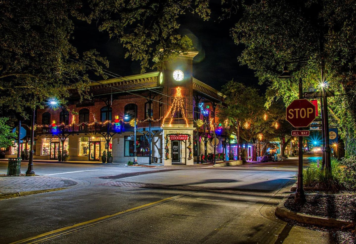 A historic building stands prominently at the intersection, with its clock tower illuminated against the night sky. Colorful lights adorn the façade, while tree branches provide a natural canopy. Street signs and lampposts enhance the charming atmosphere of the downtown area.