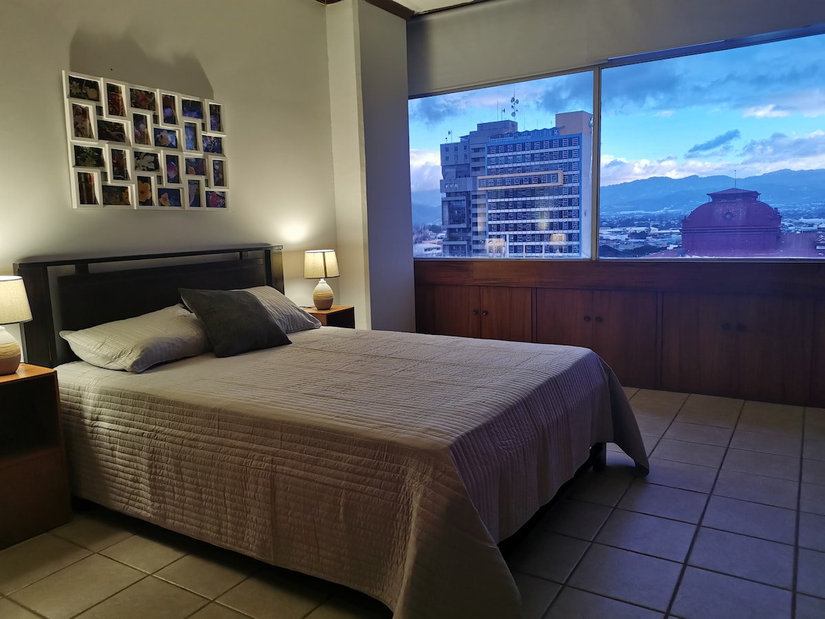 A spacious bedroom is equipped with a large bed covered with a light quilt. Bedside tables with lamps are placed on either side of the bed. A large window offers views of the city skyline, with soft lighting enhancing the room's ambience.