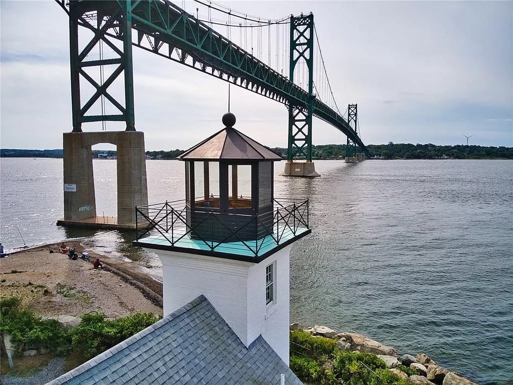 The lighthouse stands prominently at the water's edge, featuring a charming lantern room atop. A bridge spans the bay in the background, with gentle waves lapping at the shore. A pebbled beach is visible, while moments of activity can be spotted along the water.