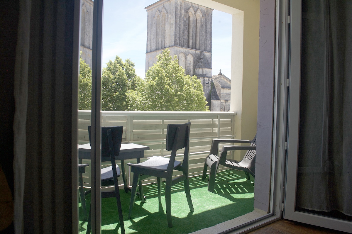 A balcony is visible through glass doors, featuring a small table and chairs on green artificial turf. The view includes tall trees and a church tower in the background, contributing to a serene outdoor setting.