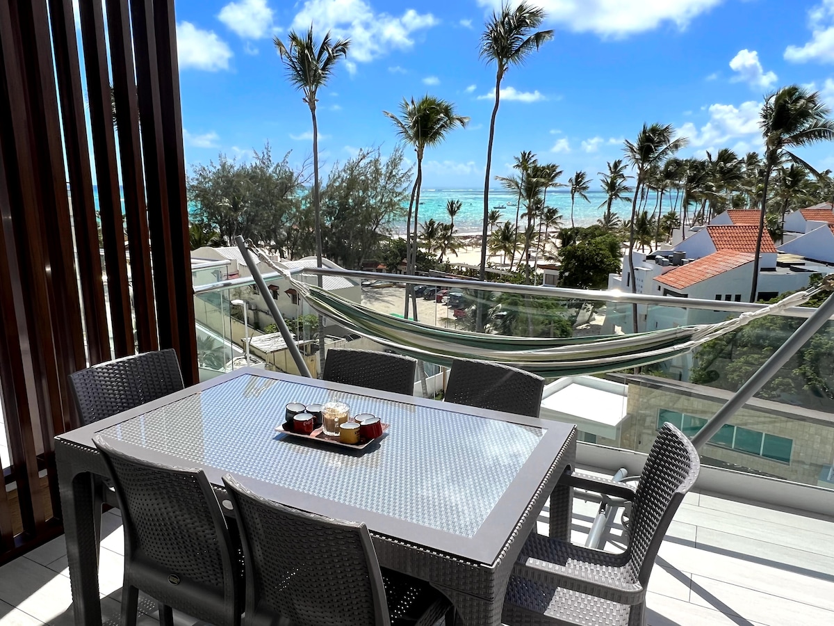 An outdoor terrace showcases a dining table surrounded by chairs, with a hammock hanging in the background. The sparkling turquoise sea and palm trees are visible beyond a glass railing, creating a scenic backdrop for outdoor gatherings.