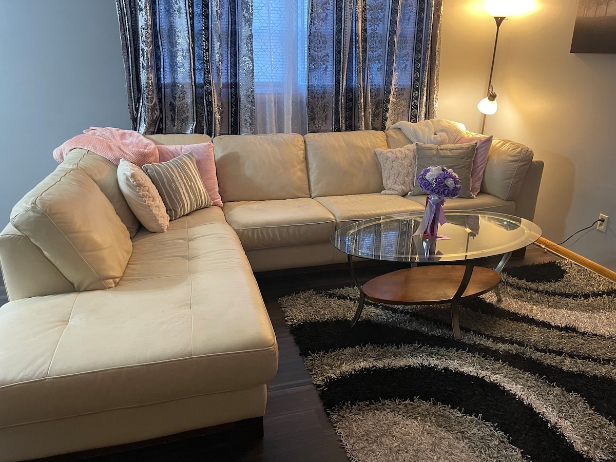 A light-colored sectional sofa is arranged in a cozy living area, accompanied by a round glass coffee table. A floral arrangement sits at the center of the table. The space features patterned curtains that frame a window, and a floor lamp adds soft lighting.