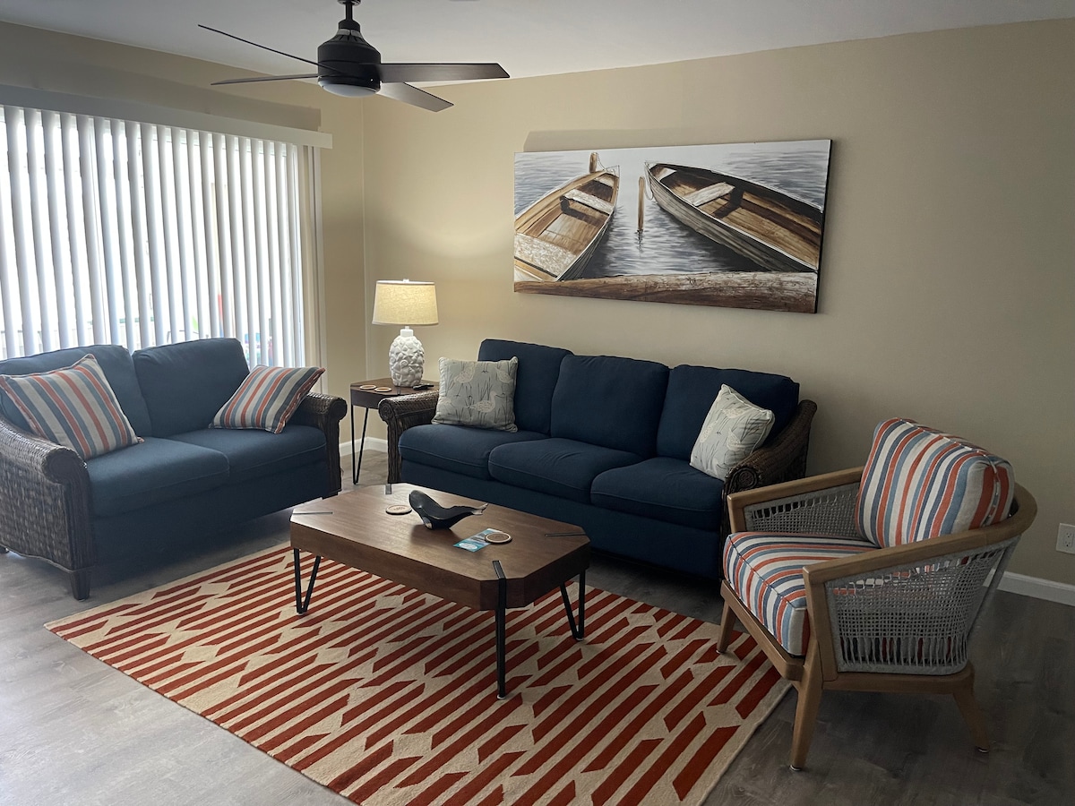 The living room features a navy blue sofa and two complementary seating options, including a patterned chair. A wooden coffee table rests on a striped area rug, while wall art depicting boat scenes adds a coastal touch. Natural light filters through vertical blinds.