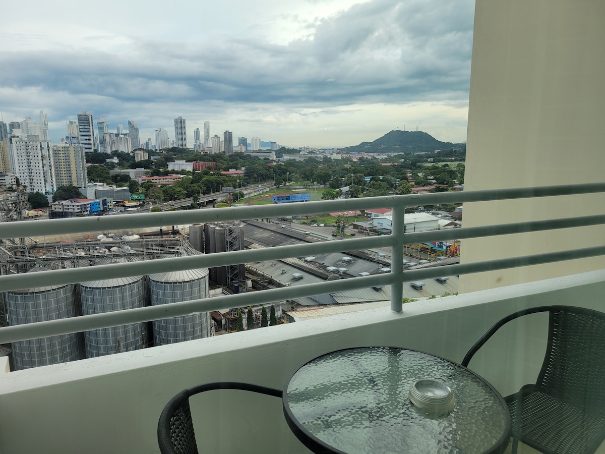 An outdoor balcony features a glass table accompanied by two wicker chairs, offering unobstructed views of the city skyline and distant hills. Large buildings and greenery are visible below, with a stretch of road and industrial facilities contributing to the bustling urban environment.