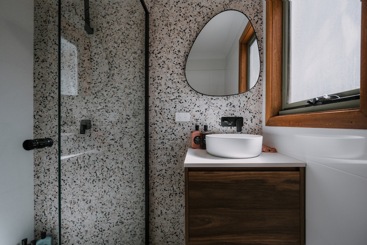 A modern bathroom features a walk-in glass rain shower with a textured wall design and a sleek, circular mirror above a white basin. Wooden cabinetry provides storage, complemented by natural light from the window.