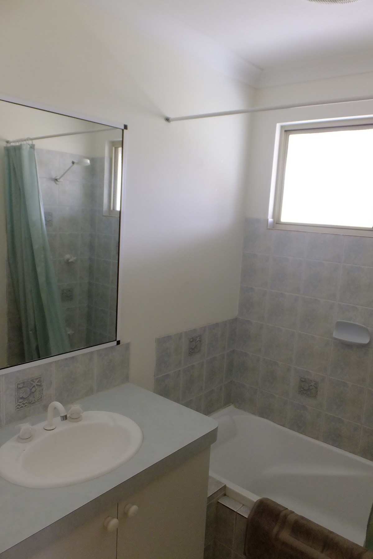 A well-lit bathroom features light-colored tiles and a combination of a shower and bathtub. A mirror occupies the wall above a simple sink with two faucets. Natural light filters in through a nearby window, enhancing the functional layout.