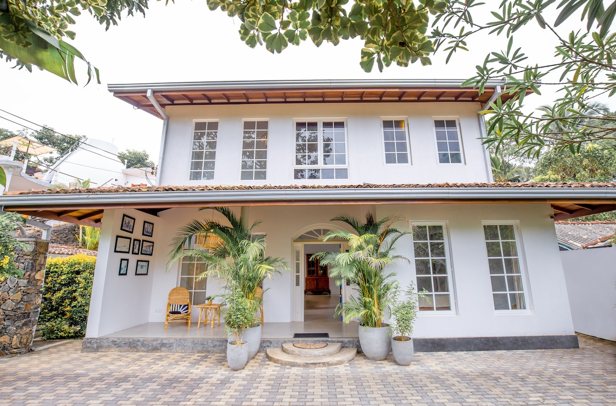 The exterior of Sunnavind House features a modern two-story structure with large windows allowing natural light. Potted plants frame the entrance, while decorative stonework and a patterned pathway lead to the inviting front door. A spacious verandah showcases seating, enhancing the home's welcoming appeal.