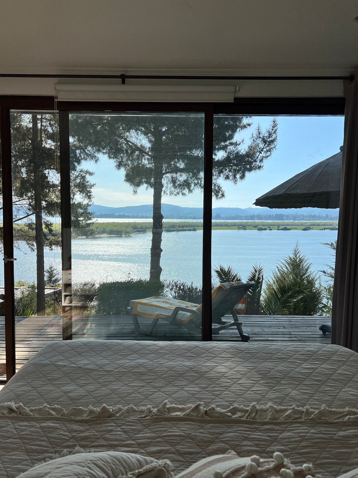 A serene view of the lake is framed by large sliding glass doors. Outside, a wooden deck is visible with a chair and an umbrella. Lush greenery and distant mountains enhance the landscape, while soft light is cast across the scene.