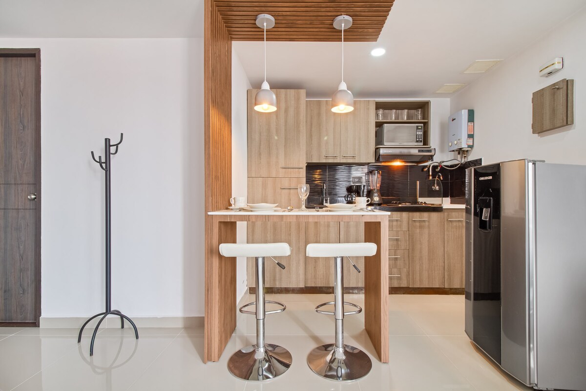 A modern kitchen area features wooden cabinetry and stainless steel appliances, including a microwave and refrigerator. Two white bar stools are positioned at a small countertop, creating a cozy dining space. Natural light illuminates the area, enhancing the neutral color palette of the studio.
