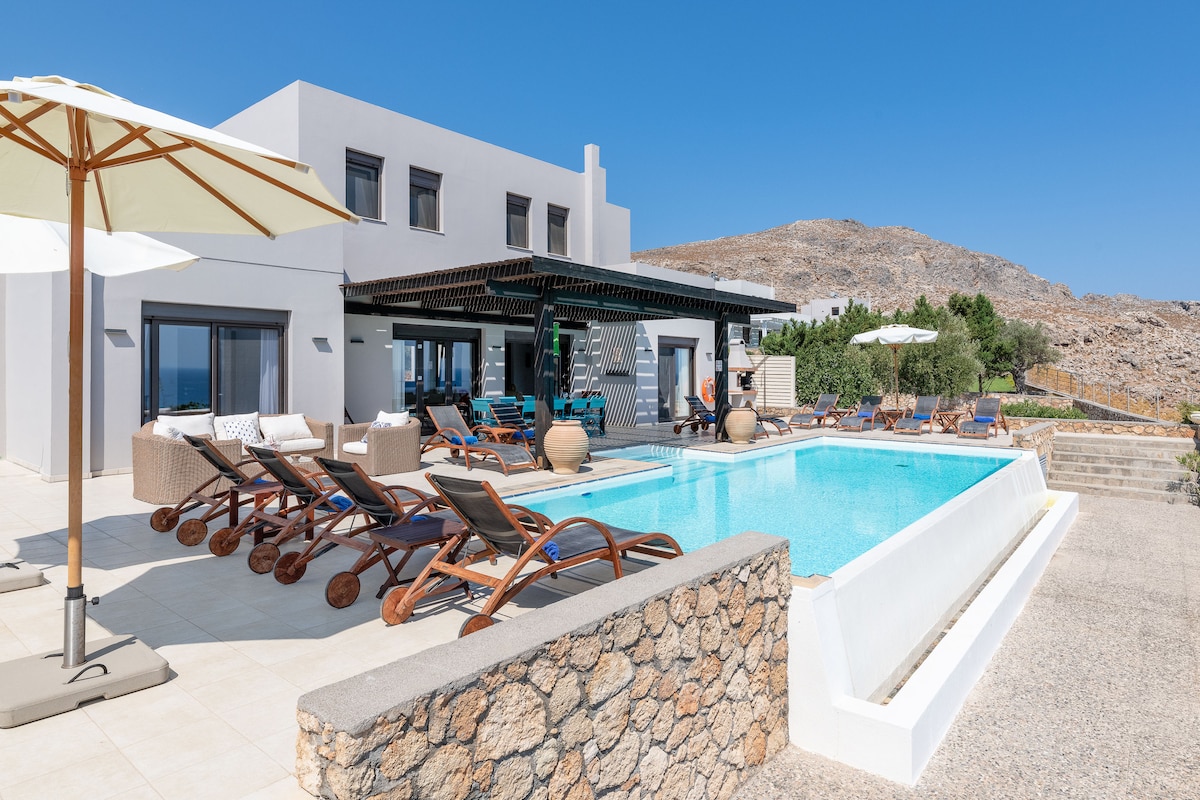 An outdoor area features a private pool surrounded by sun loungers and umbrellas. Natural stone walls enclose the space, and panoramic views of the mountainous terrain and sea are visible in the background. Various lounge seating options are arranged around the pool for relaxation.