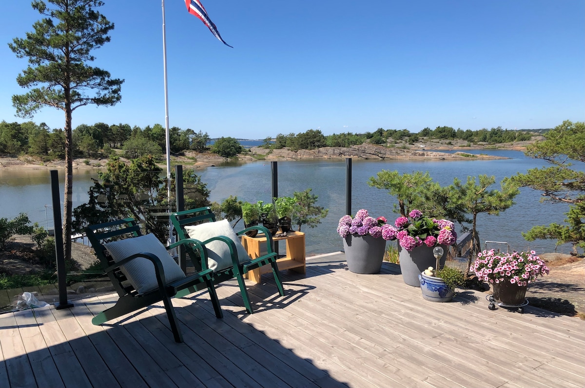 A sunlit terrace features two green chairs overlooking a serene water view. Potted flowers in various colors adorn the edges, while a small table is positioned nearby. The surrounding landscape includes trees and rocky formations under a clear blue sky.
