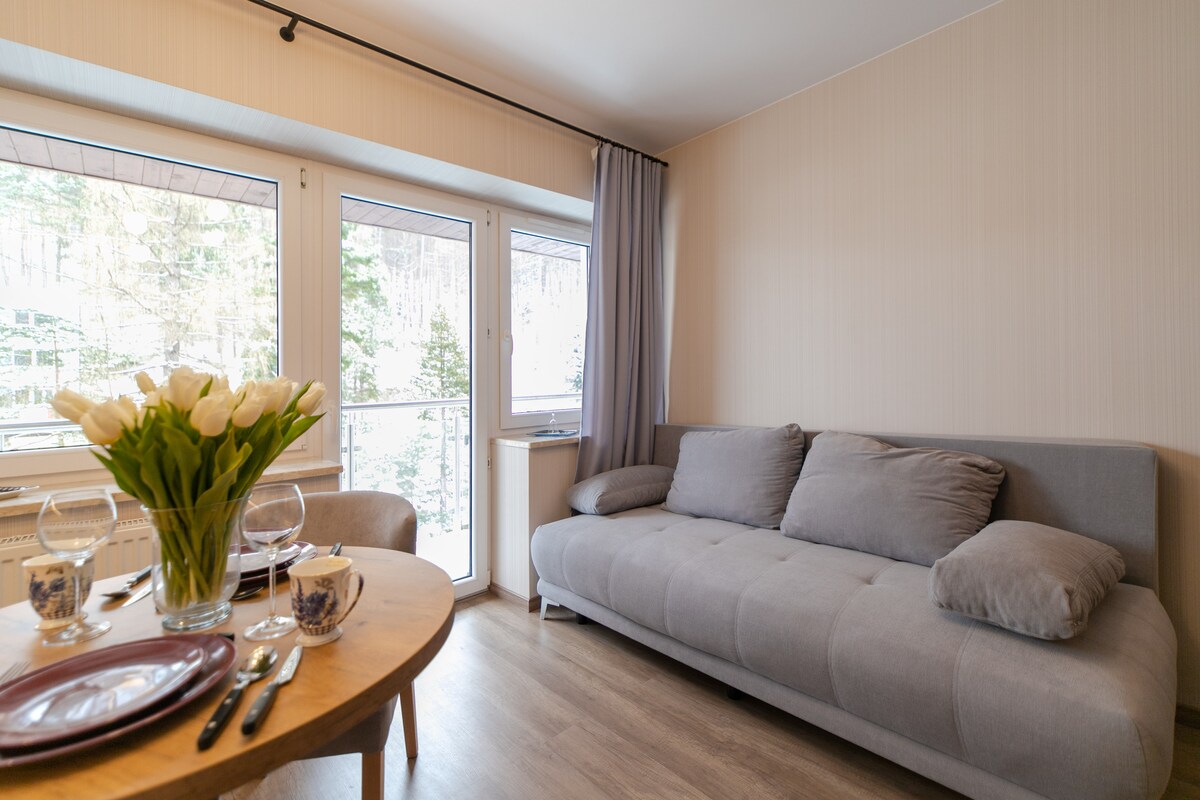 A cozy seating area is presented with a light gray sofa positioned near large windows, allowing natural light to fill the room. A round wooden dining table is set with elegant tableware, complemented by a vase of fresh tulips, enhancing the warm atmosphere.