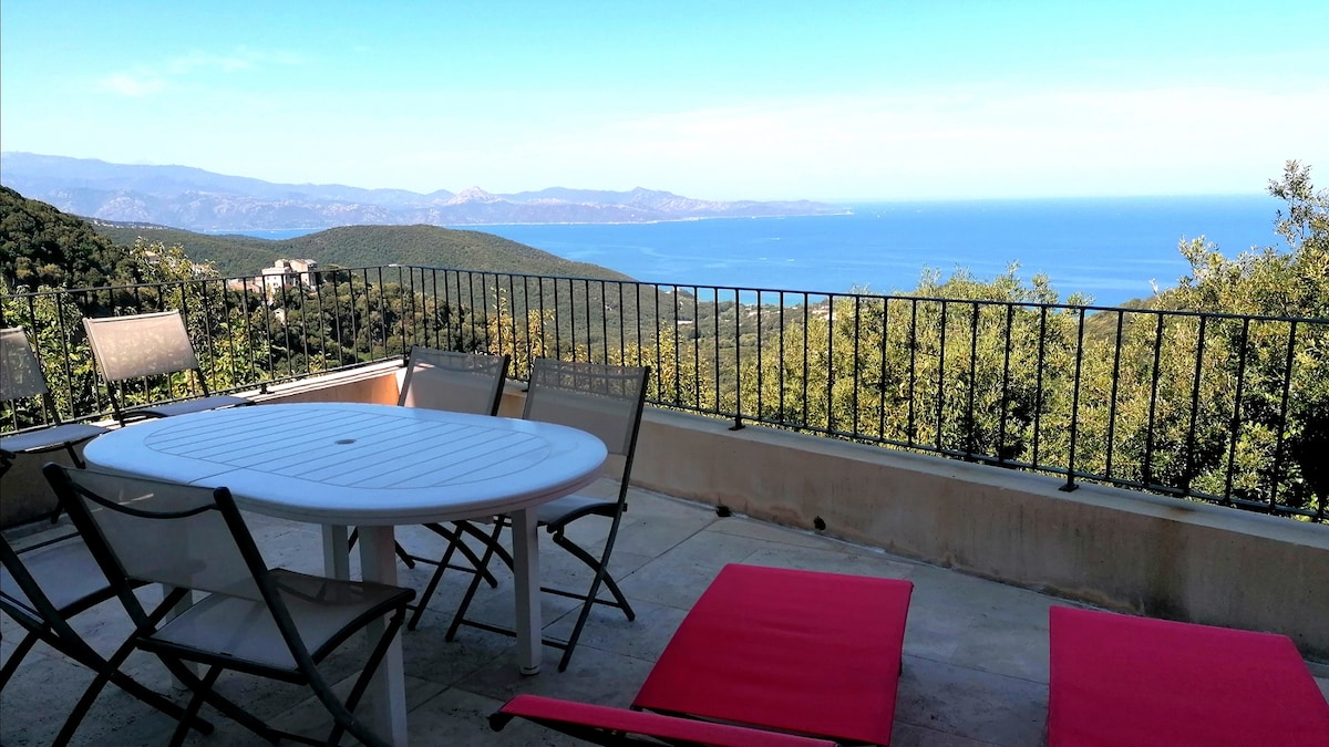 An outdoor terrace area features a round table surrounded by six folding chairs, complemented by two lounge chairs. A panoramic view of the sea and distant mountains is visible, showcasing a bright blue sky. The terrace is enclosed by a low iron railing.