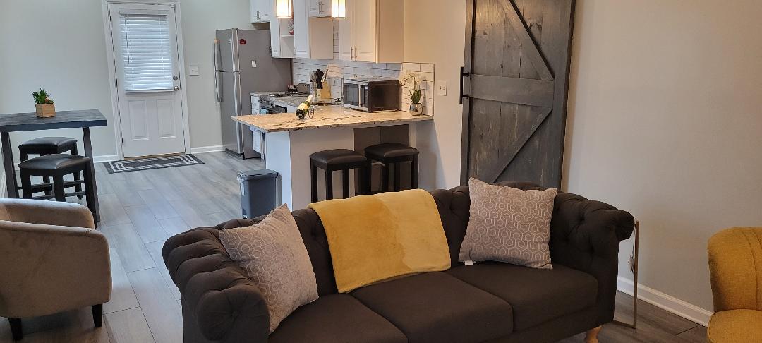 The living area features a comfortable dark couch with decorative pillows, arranged towards a modern kitchen with light wood cabinets and a granite countertop. A dining table is positioned nearby, and a rustic barn door adds character to the space. Soft lighting enhances the inviting atmosphere.