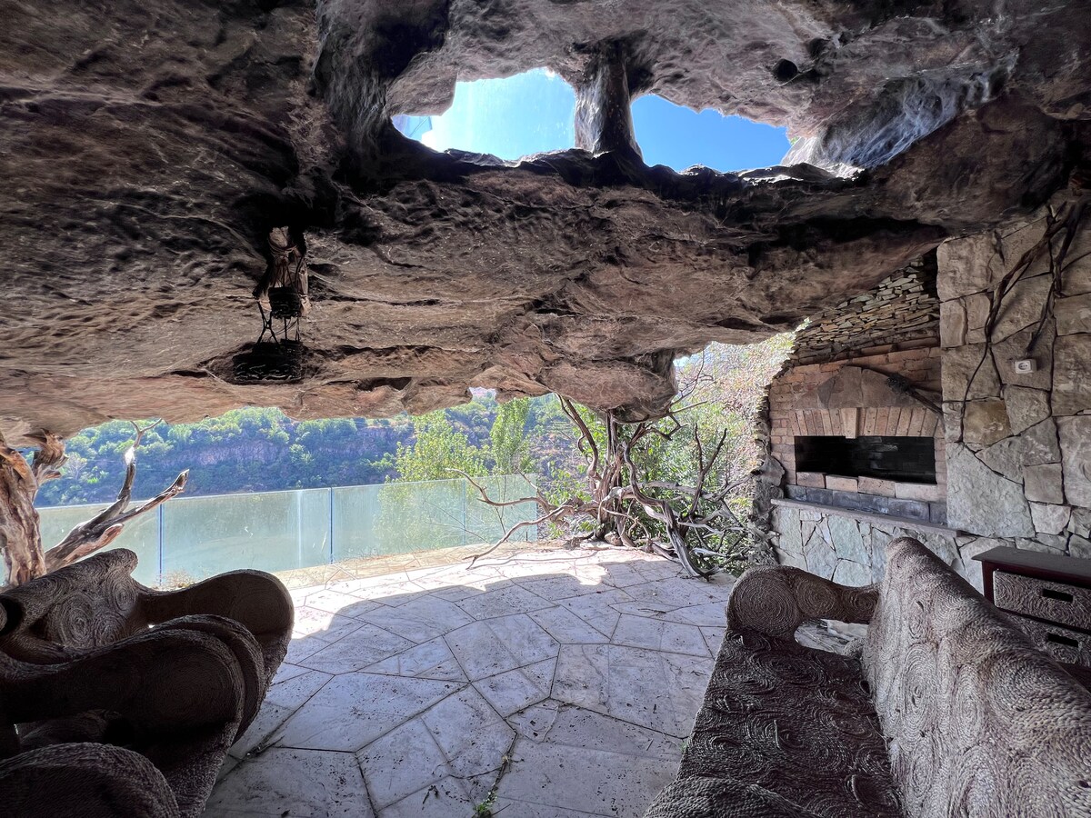 A unique seating area is situated under a natural stone overhang, featuring textured furniture arranged for relaxation. Large openings above allow natural light and views of the surrounding landscape, with a stone structure suggesting an outdoor cooking area nearby.