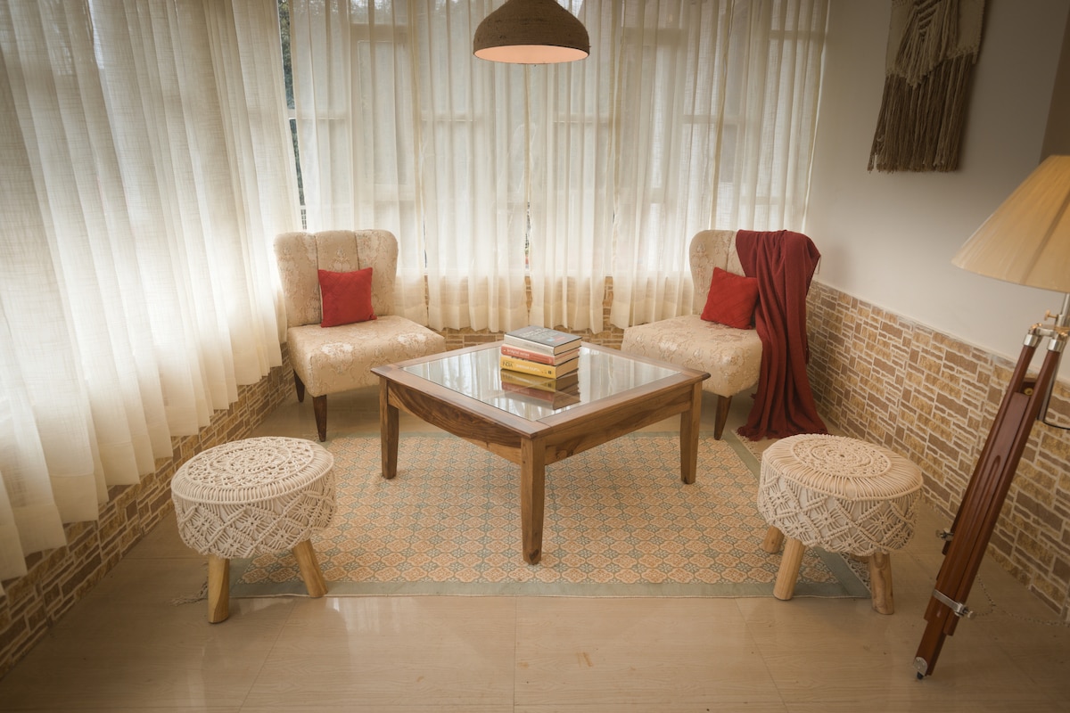 A cozy sitting area is created with two plush armchairs and a wooden coffee table. Light streams through large windows covered with sheer curtains, illuminating the space. Decorative poufs and a rich textile accentuate the neutral tones of the room.