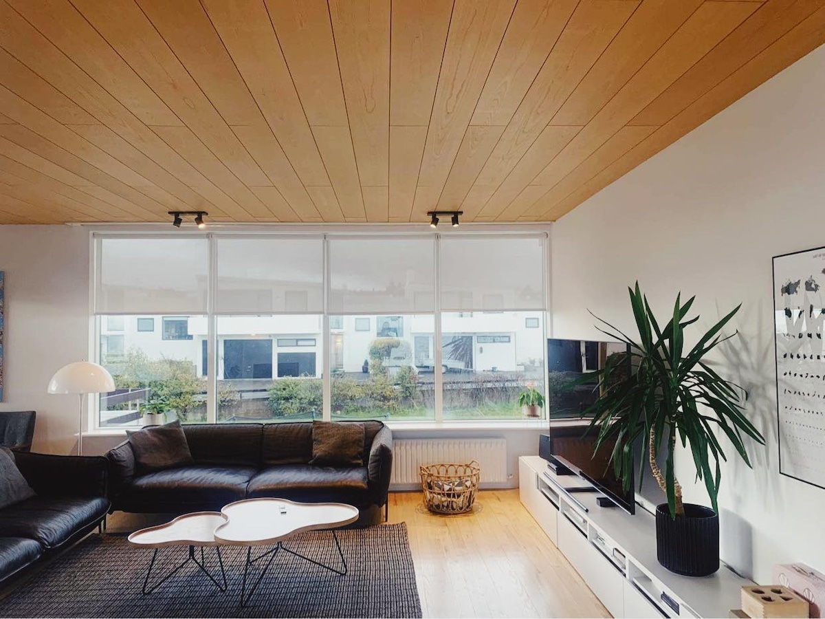 A spacious living room features large windows that provide ample natural light. A modern black leather sectional sofa is positioned around a unique coffee table, with a minimalist TV unit and decorative plant completing the inviting atmosphere.