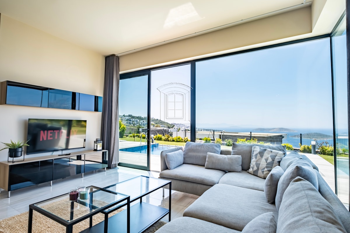 A spacious living area features large glass doors offering views of the outdoor pool and landscape. A comfortable sectional sofa is arranged around a modern coffee table, with a television mounted on the wall. Natural light fills the room, enhancing its airy atmosphere.