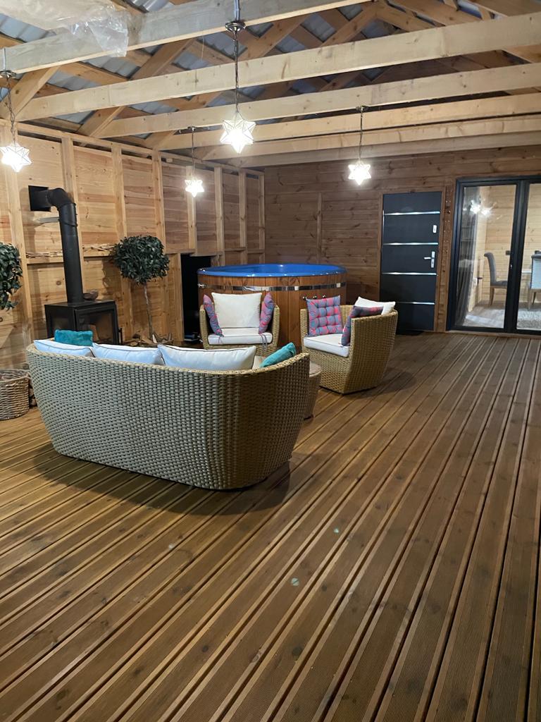 A cozy seating area features two woven chairs with colorful cushions, arranged around a circular bar. Wooden walls and flooring create a rustic feel, while pendant lights hang from the ceiling, offering a warm glow. Large glass doors provide access to an outdoor space.