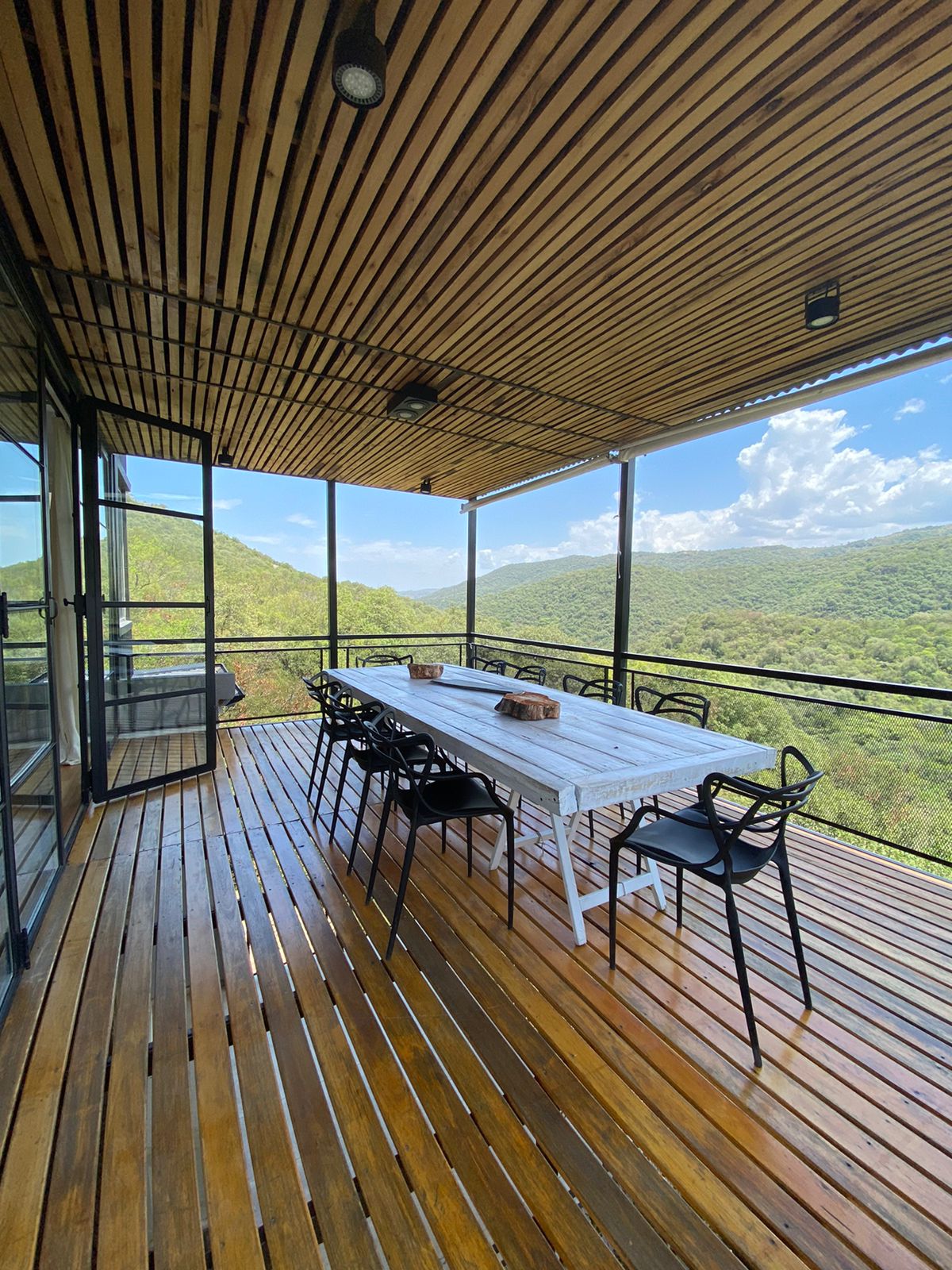 A covered outdoor deck is present, showcasing a long dining table surrounded by black chairs. The natural wood flooring complements the surrounding mountain views visible through large glass panels. Bright sky and green hills create an inviting setting for outdoor dining.