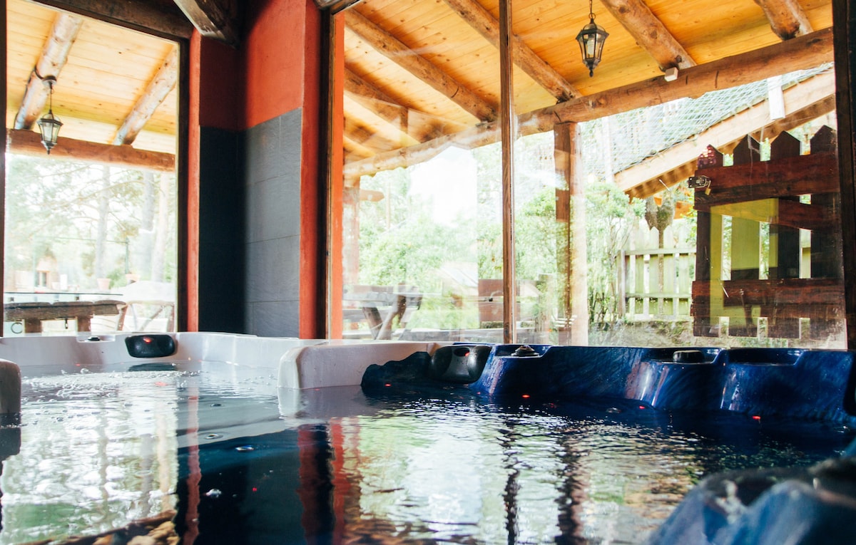 The image showcases a spacious indoor hot tub area, featuring two large tubs filled with clear water. Large glass windows provide a view of the natural surroundings, allowing natural light to illuminate the wooden beams and rustic decor of the room.