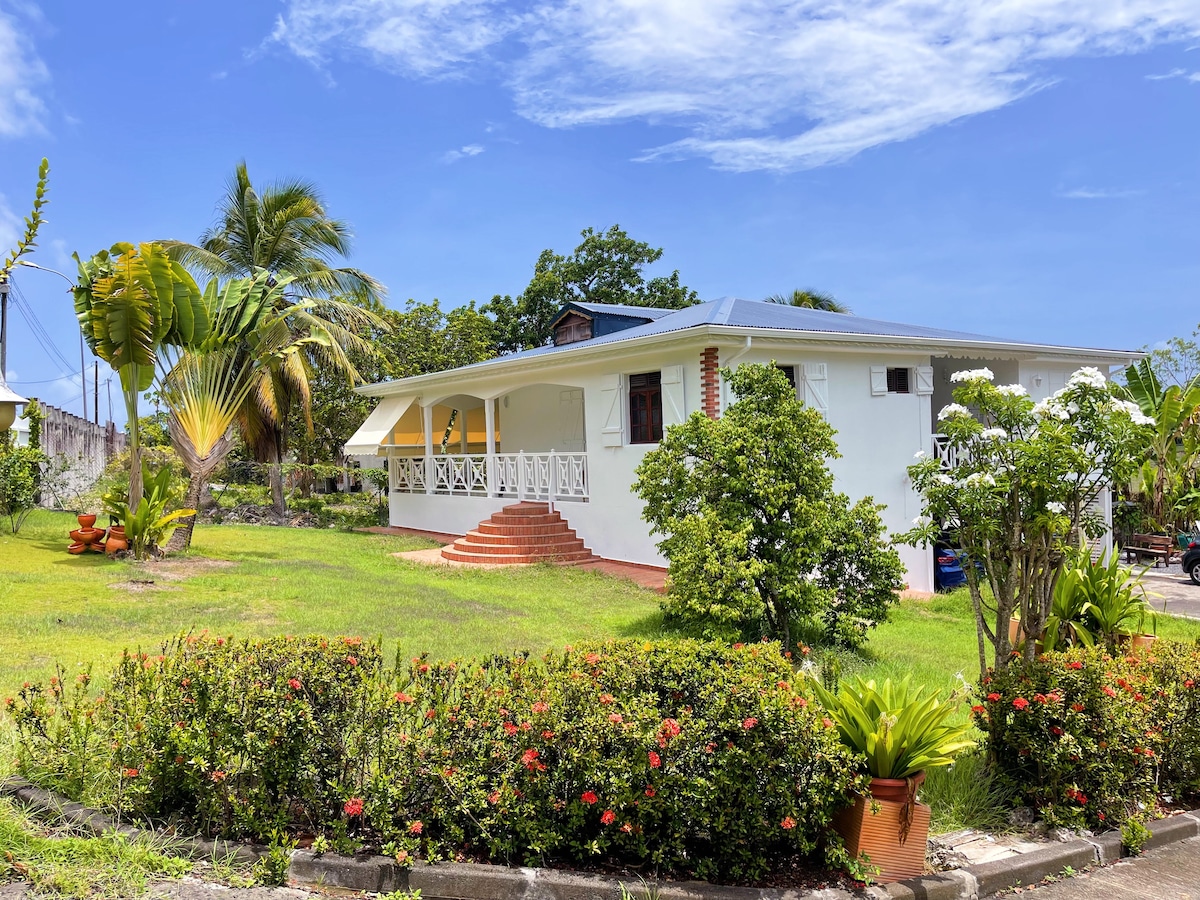 A bright, single-story villa is surrounded by lush greenery and colorful landscaping. The structure features a covered porch with white railings, providing a view of the garden. Palm trees and flowering plants enhance the serene outdoor setting under a clear blue sky.