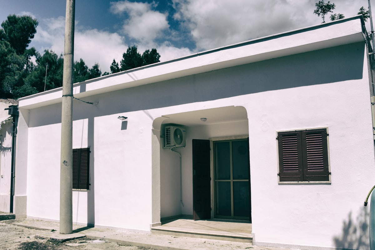 The exterior of a single-story house displays a clean white facade, featuring dark wooden shutters and a glass door leading to the entrance. An air conditioning unit is mounted on the wall, contributing to the comfort of the space. A gravel pathway is visible.