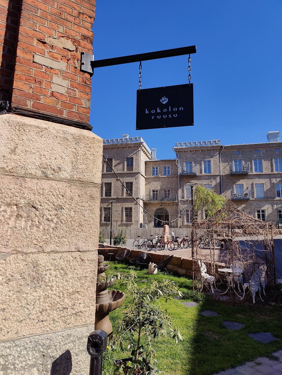 A sign for 'kakolan ruusu' is seen hanging from the building, surrounded by a well-kept outdoor area featuring greenery and a stone pathway. Historic architecture is visible in the background, creating a charming atmosphere.