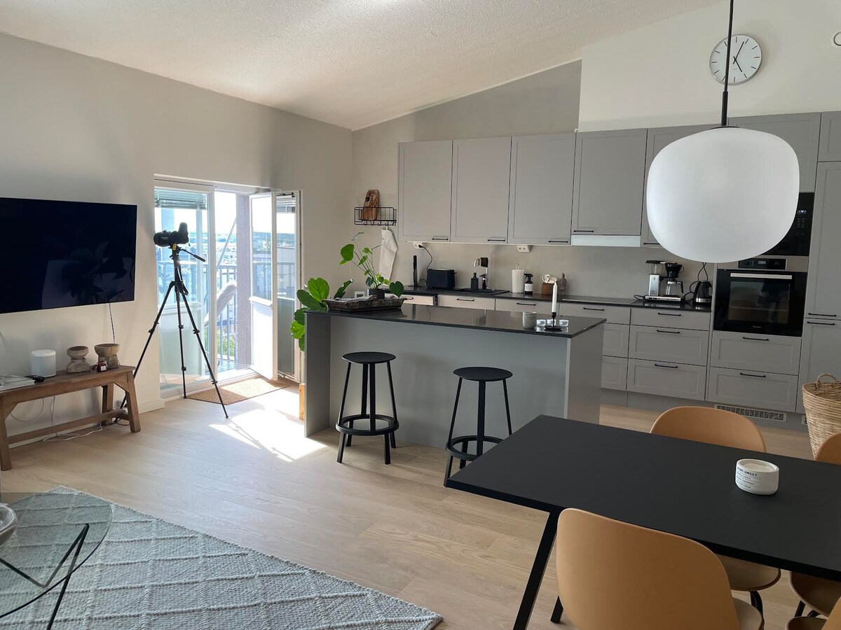 A modern kitchen area features a central island with seating for two. Gray cabinetry complements a sleek black countertop. Sunlight streams through large glass doors that open to a balcony. A comfortable dining table and a cozy seating arrangement are visible in the open space.