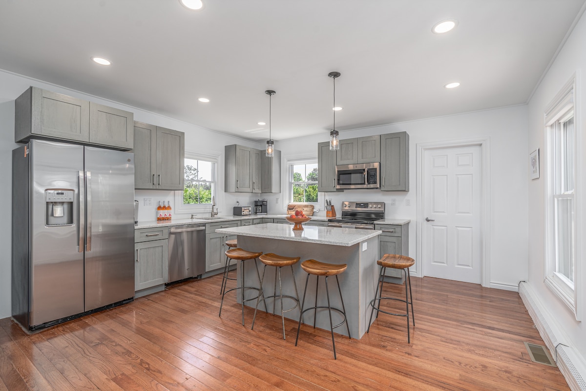 A modern kitchen features light gray cabinets and stainless steel appliances. A central island with three wooden stools offers additional seating. Soft lighting illuminates the space, while large windows provide views of the surrounding greenery, enhancing the kitchen's bright and airy feel.