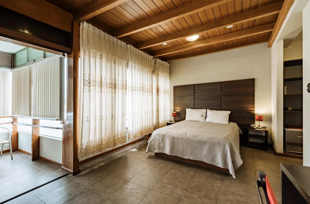 A spacious bedroom features wooden ceiling beams and large windows covered with sheer and adjustable blinds. A neatly made bed, flanked by bedside tables, is complemented by soft lighting. Neutral tones are present in the decor, creating a calm and inviting space.