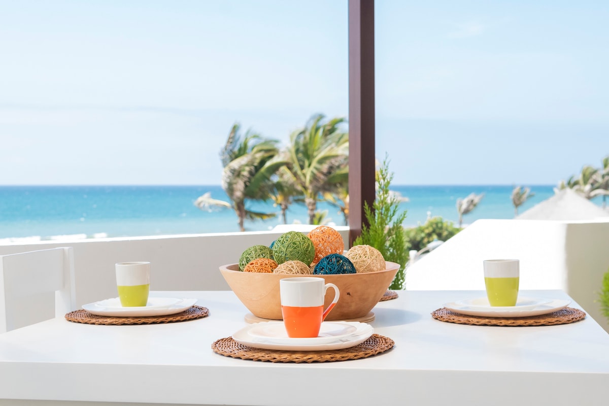 An outdoor dining area is presented with a white table set for three, featuring colored cups and woven placemats. A bowl filled with decorative spheres sits at the center, with a backdrop of the ocean and swaying palm trees creating a tranquil setting.