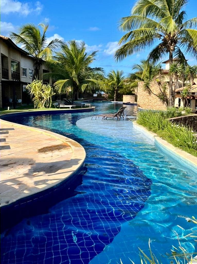 A large, winding pool is surrounded by tropical palm trees and lush greenery. Lounge chairs are positioned along the water's edge. The pool reflects clear blue skies, creating a serene atmosphere for relaxation. Buildings are visible in the background, contributing to the resort-like setting.