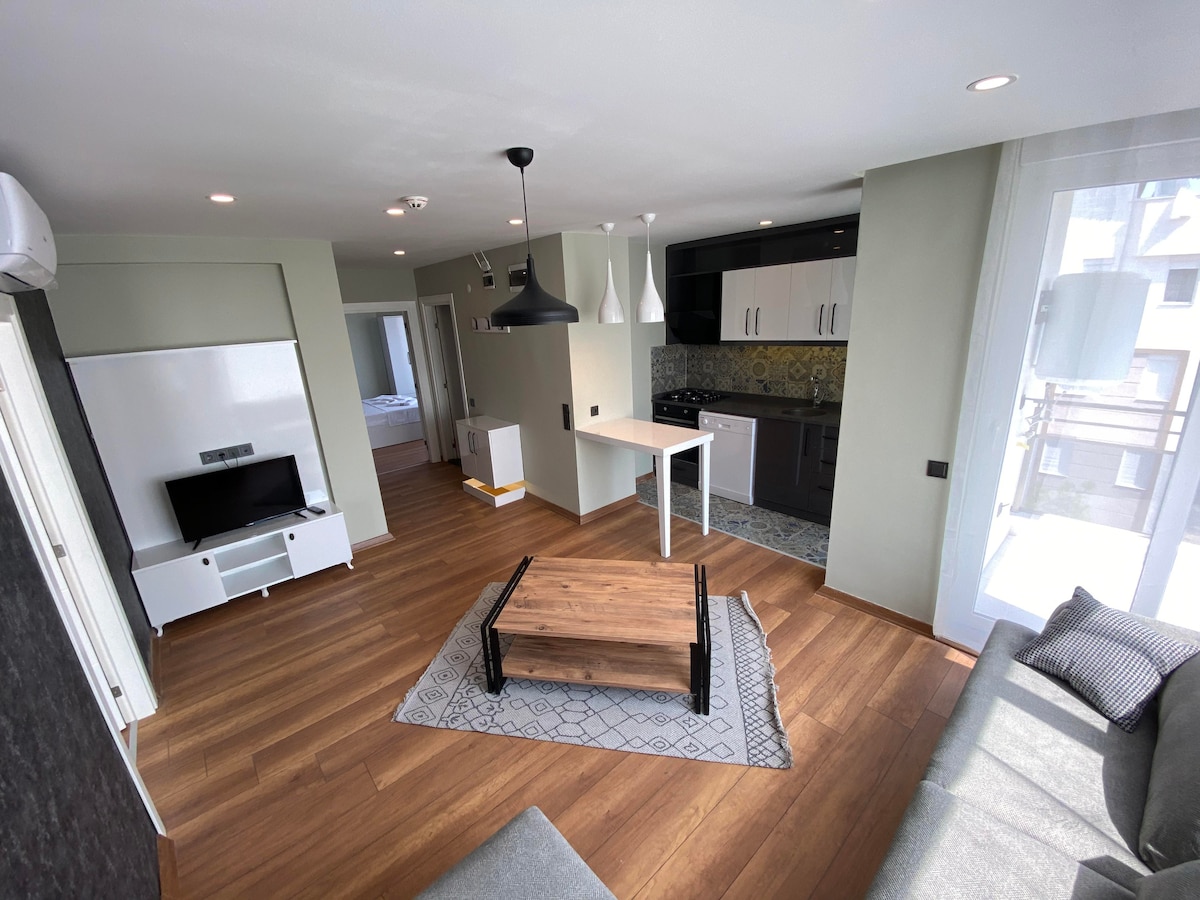 An open living space is presented, featuring a modern sofa and a coffee table that contrasts with the dark wood floor. A sleek kitchen area lies in the background, with black cabinetry and decorative tiles visible. Natural light filters in through large windows, enhancing the inviting ambiance.