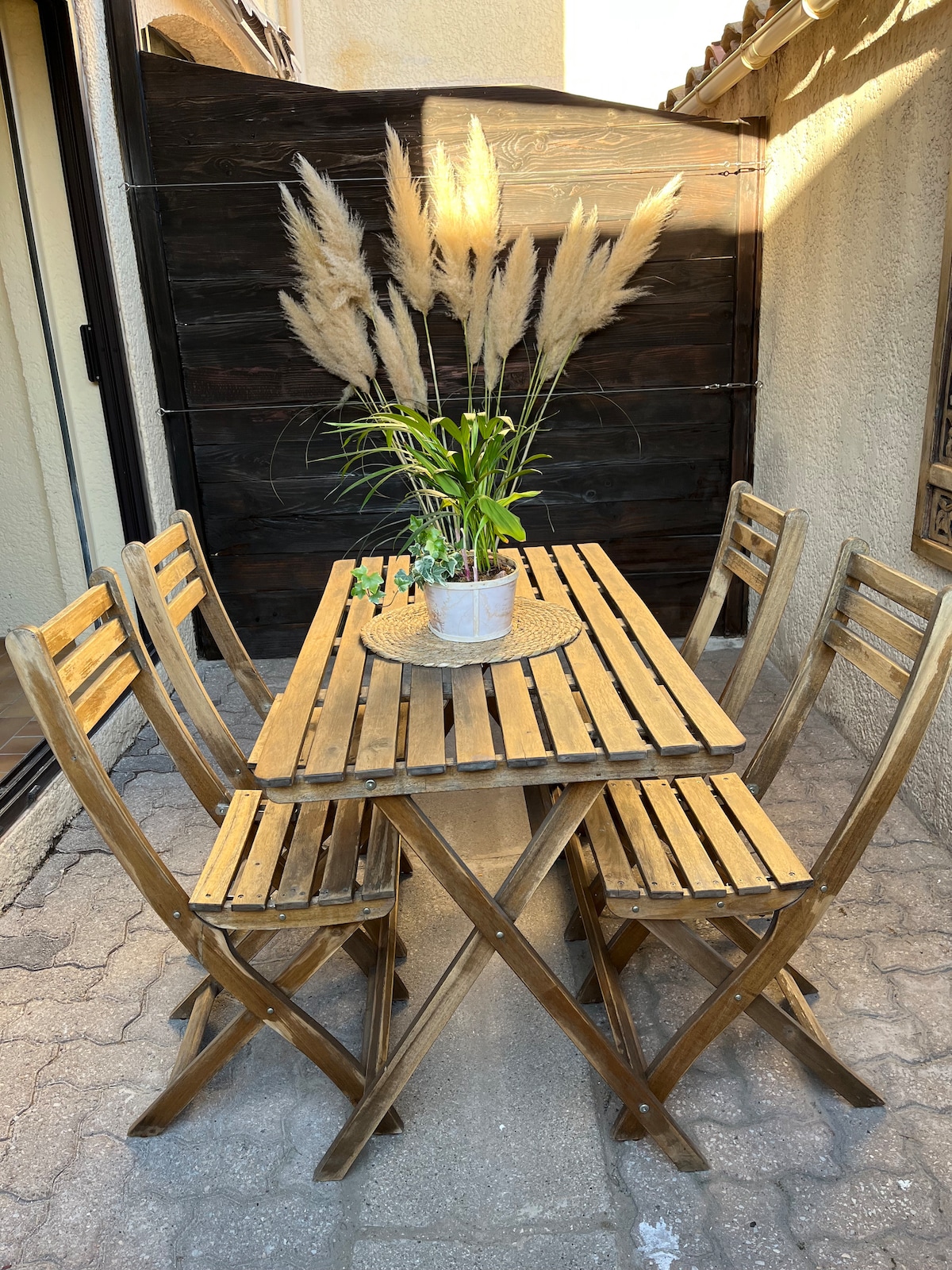 A wooden outdoor dining table is surrounded by four matching folding chairs. A potted plant sits in the center on a round tray, adding a touch of greenery to the cozy terrace space.
