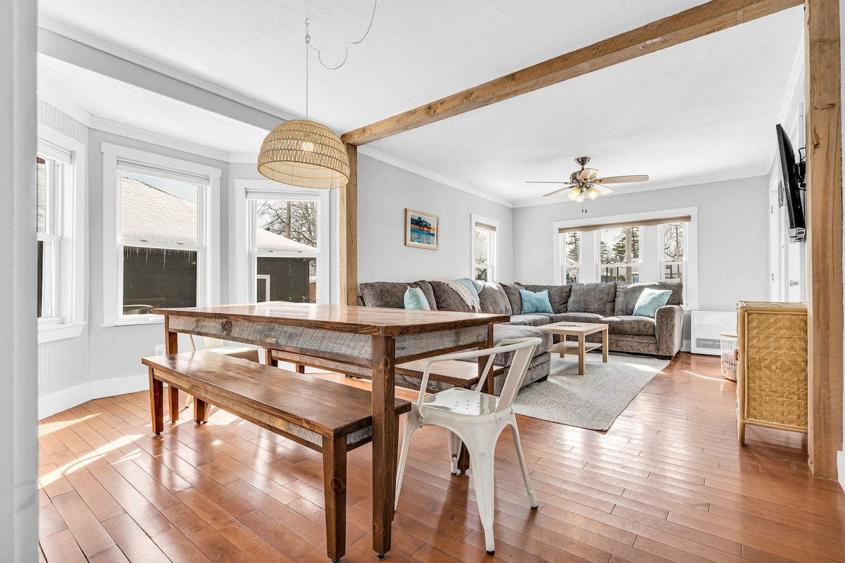 A bright and airy living space features a large sectional couch and a dining table made of natural wood. Sunlight streams through multiple windows, illuminating the warm wood flooring. A ceiling fan and wall-mounted TV provide comfort and entertainment in this welcoming area.