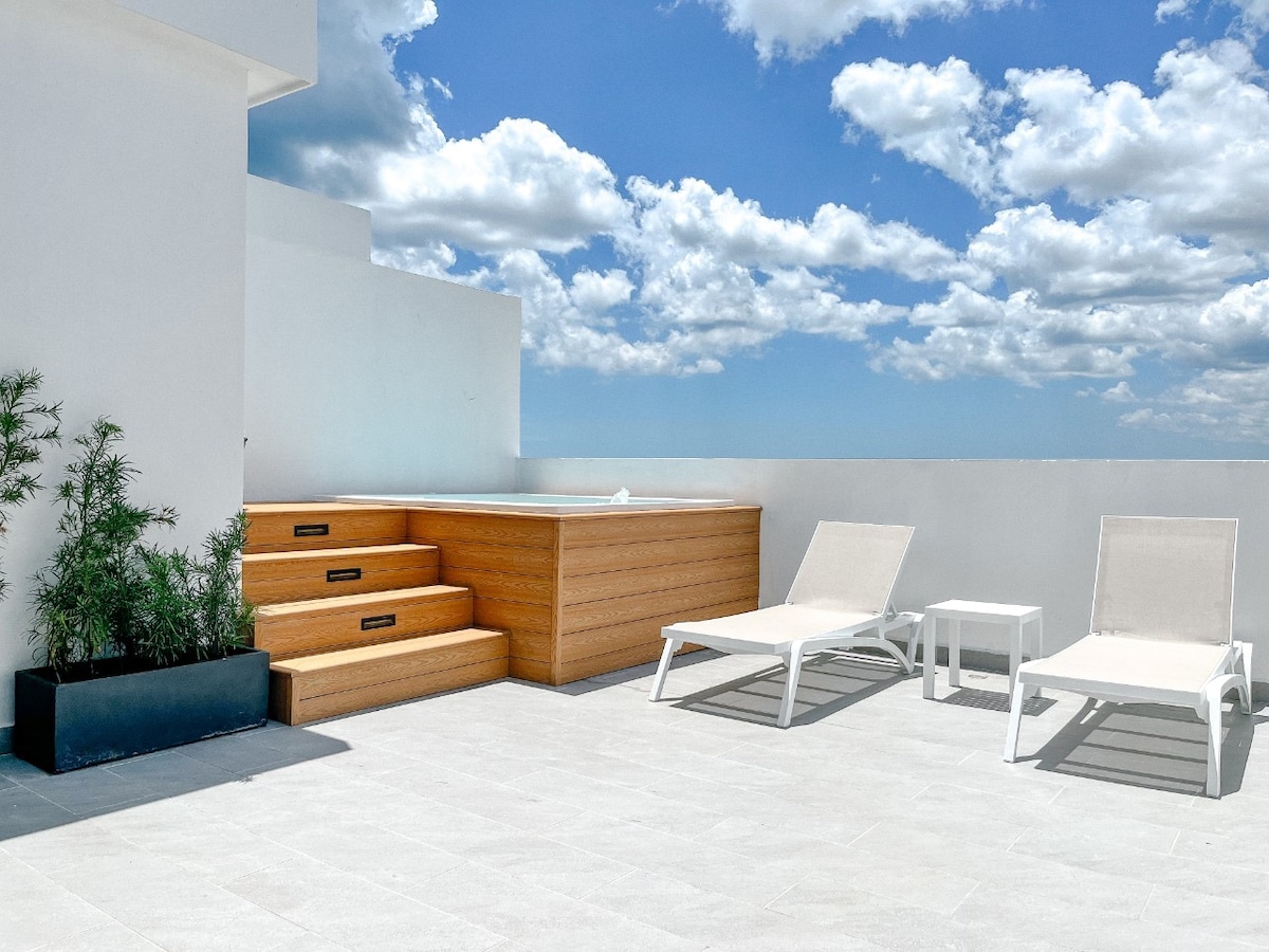 A private terrace features a jacuzzi, surrounded by stairs leading up to it. Two sun loungers and a small table are positioned on the light-colored tile floor, with greenery in a black planter adding a natural touch. The sky is bright with scattered clouds.