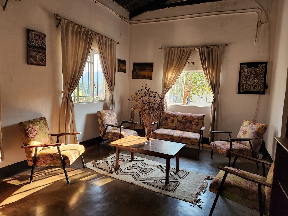 A cozy living area is furnished with two floral-patterned sofas and four armchairs. A wooden coffee table sits atop a woven rug, while sunlight filters through the large windows dressed in light curtains, creating a warm ambiance.