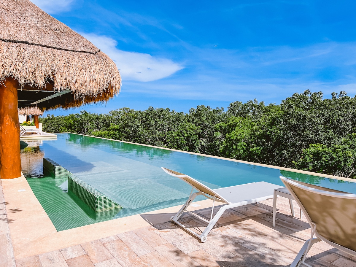 An infinity pool extends over the edge, framed by lush greenery. Sun loungers are placed along the poolside, providing a spot for relaxation. A shaded area features a thatched roof, enhancing the tranquil setting.