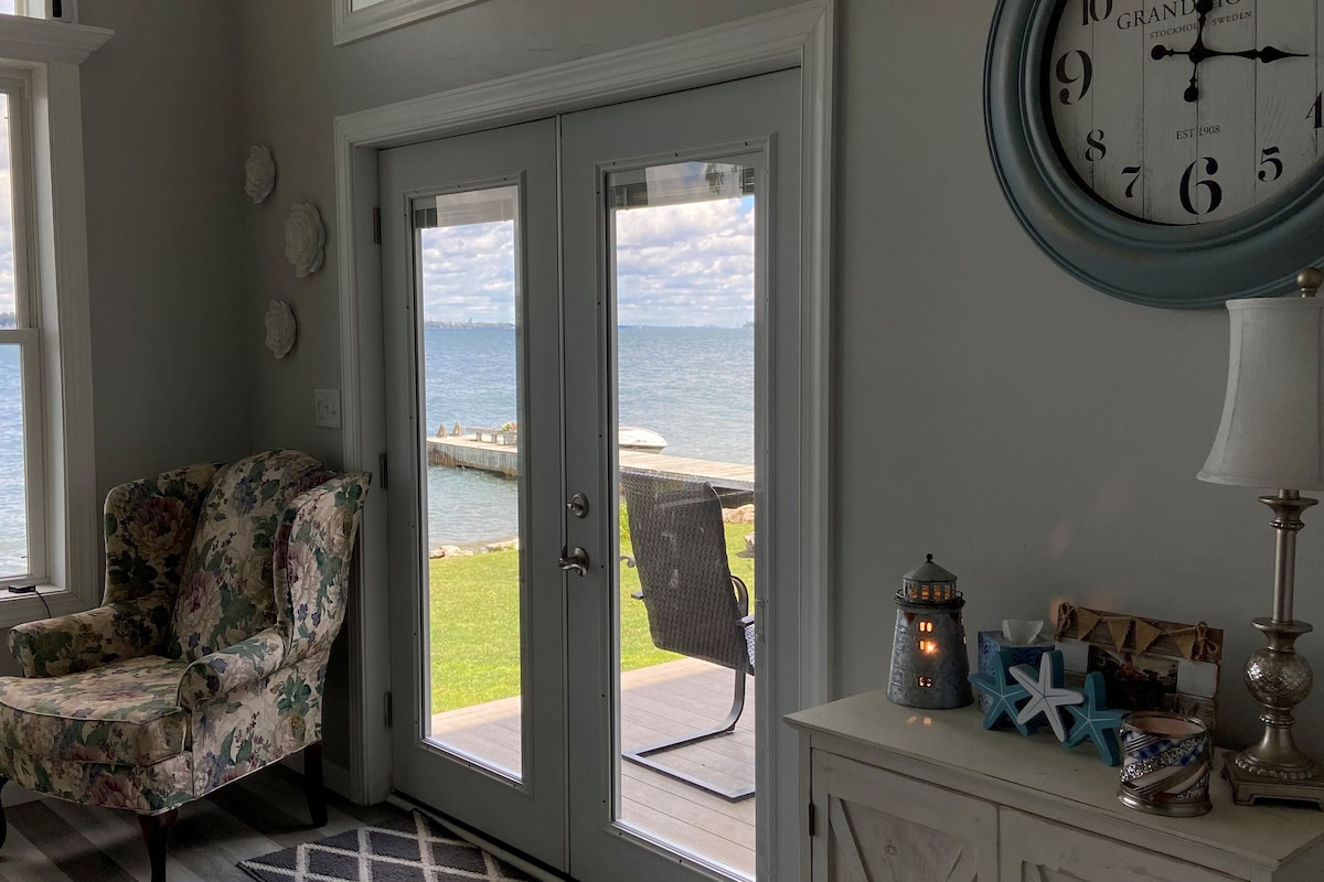 Large glass doors frame a scenic view of the St. Lawrence River, allowing natural light to fill the space. An inviting floral armchair and a decorative clock enhance the interior. The outdoor area features a private dock extending towards the water, complemented by a peaceful grassy shoreline.